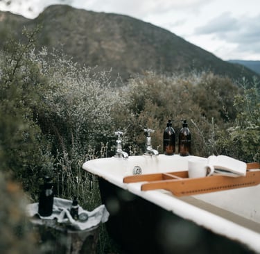An outdoor clawfoot bathtub with a wooden tray and toiletries set against a scenic mountain landscape.