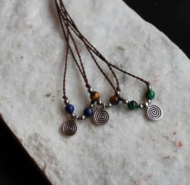 Drei Ketten mit braunem Garn und einem silberfarbenen Spiralenanhänger. Jede Kette hat andere Perlen: blaue, grüne oder braun