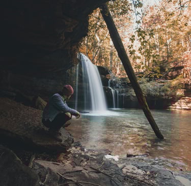 Person resting by waterfall with backpack, showing natural shelter and wilderness hydration