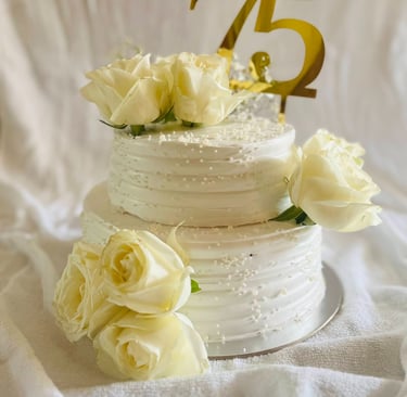 Two-tier white buttercream 75th birthday cake decorated with fresh white roses and a gold topper.