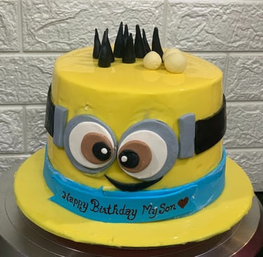 Yellow Minion birthday cake for a son with goggles and a blue ribbon banner on a cake stand.