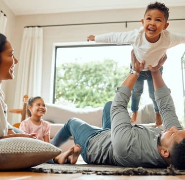 Una familia está jugando en la alfombra, el niño está sostenido en el aire por su padre.