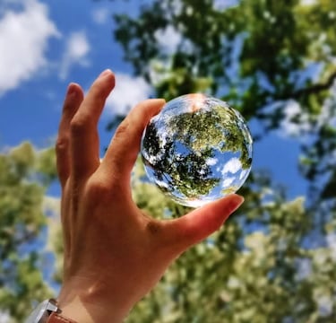 A hand holds a small glass globe reflecting trees and sky, symbolizing the beauty of creation.