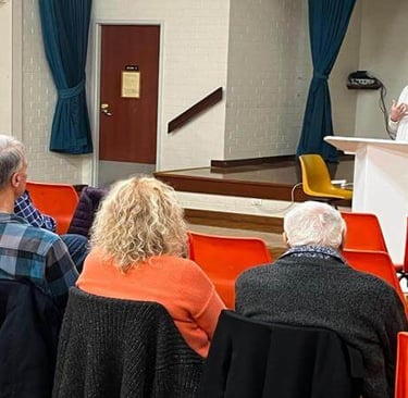 Candice speaks at a podium to a small seated audience in a community hall during a church gathering.