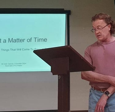 Collin stands at a lectern reading from notes while a presentation titled “It’s Just a Matter of Tim