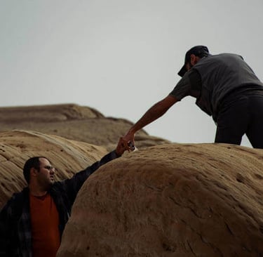 One person reaching down to help another climb a rocky slope under a cloudy sky