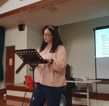 Hazel reads from a stand during a church meeting, with a projected prayer or declaration visible in 