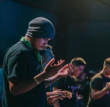 Group of people in dim light with eyes closed and hands raised in reflective or prayerful posture.