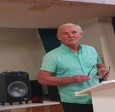 Len stands at a podium holding glasses, smiling as he addresses a church group in a relaxed setting.