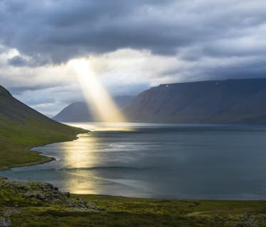 Sunlight breaks through clouds over a peaceful valley and water, showing divine presence in nature.