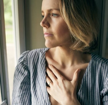 A woman gazes out a window with her hand on her chest, suggesting reflection and longing.