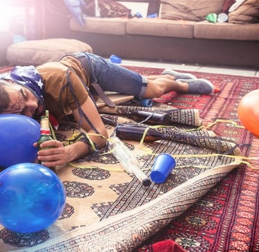 A man sleeping on a rug in a messy room after a house party with balloons and empty beer bottles.