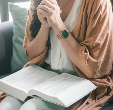 eated person with clasped hands and open Bible on lap, bathed in soft indoor light.