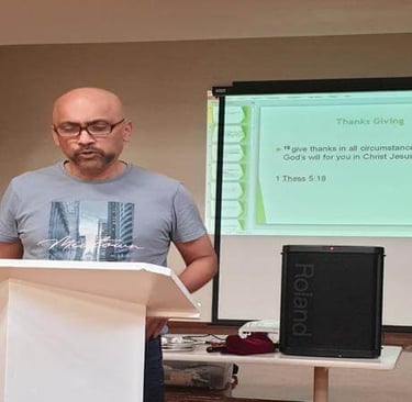 Raju reads from a lectern during a church gathering, with a slide behind him displaying a Bible vers