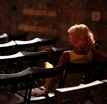 Person in yellow dress seated alone among empty chairs in a softly lit room.