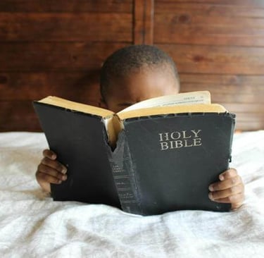 Young child holding a large black Bible, seated on a bed with wooden paneling behind.