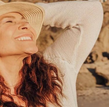 Person with long hair and woven hat smiling with eyes closed, standing near a sunlit rocky shore.