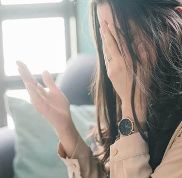 Person seated indoors with hand covering face and other hand raised, lit by window light.