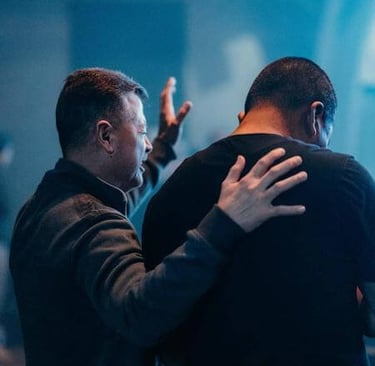 One person praying with a hand on another’s shoulder, symbolizing support, prayer, and community.