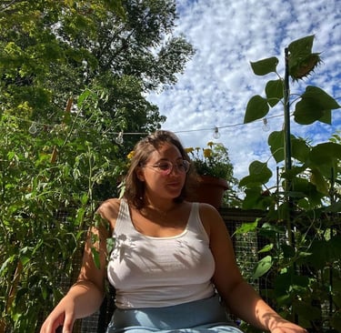 A woman wearing glasses and a tank top sits in a lush garden under a blue sky with clouds.