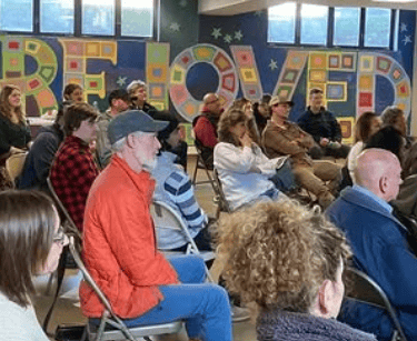 A diverse group of people sitting in a community room for a presentation in front of a colorful mural.