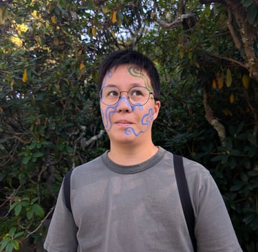 Person with blue and green artistic face paint patterns posing outdoors in front of leafy trees.