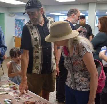 A group of people gathering around a table to look at a collection of small art cards at a community event.