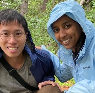 A smiling young man and woman wearing rain jackets while hiking in a lush green forest.
