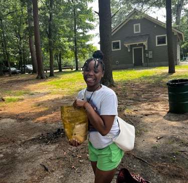 A smiling young woman holding a snack bag at an outdoor park with a cabin and tall pine trees.