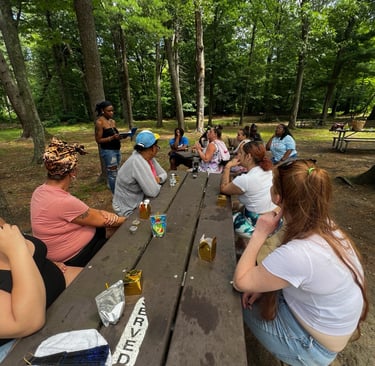 A diverse group of women sitting at a picnic table for an outdoor gathering in a wooded park.