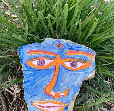 Hand-painted rock art featuring a blue face with orange accents nestled in green grass.
