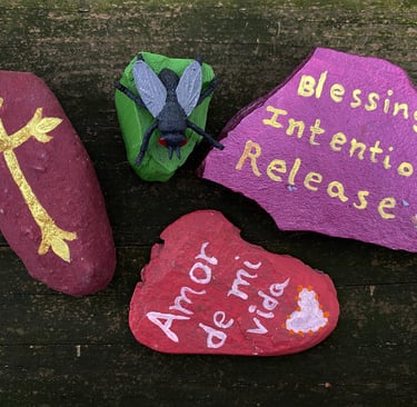 Hand-painted stones with a gold cross, fly figurine, and spiritual messages on a rustic wood background.