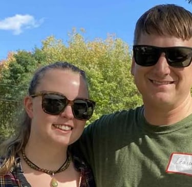 A smiling couple wearing sunglasses posing outdoors on a sunny day with autumn trees in the background.