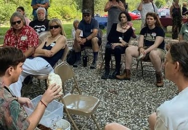 A group of people attending an outdoor educational workshop sitting in a circle on gravel.