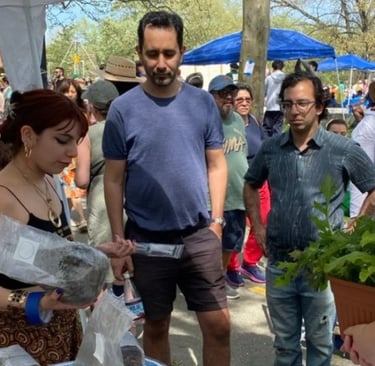 Customers shopping for organic plants and local goods at a crowded outdoor community farmers market.
