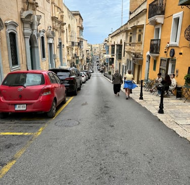 A high-angle view of a narrow, sloping street in Malta, lined with historic buildings and a church o
