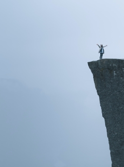 Woman standing on a tall cliff with her hands spread wide. Is it a photoshoot or will she jump?