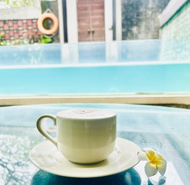 A white cappuccino cup on a glass table by a luxury swimming pool at a tropical villa resort.
