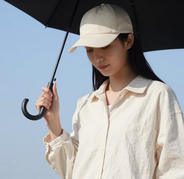 A woman wearing a beige linen shirt and baseball cap holding a black umbrella against a blue sky.