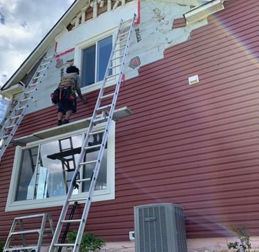 A contractor on a ladder installing red vinyl siding on the exterior of a residential home.