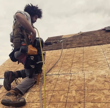 Professional roofer using a pneumatic nail gun to install plywood sheathing on a residential roof.
