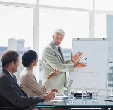 A man in a suit presents a rising graph on a flip chart to two colleagues in a bright conference room.