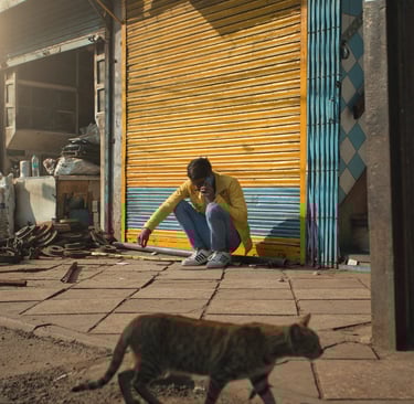 Man squatting outside a closed shop as a cat walks past in Old Delhi