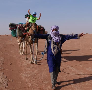 A desert guide in traditional dress leads two camels in the Sahara carrying children