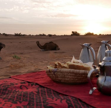 Teapots and a basket of bread laid out for tea in the desert, as two camels look on