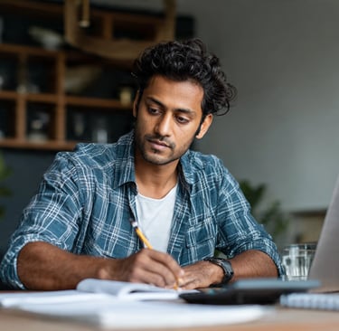 A focused man working from home using a laptop and writing in a notebook at a desk.