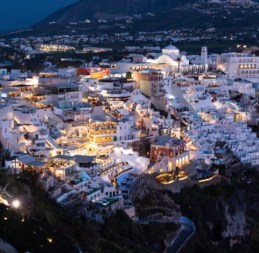 Night view of illuminated white buildings and cliffs in Fira, Santorini, Greece overlooking the Aegean Sea.