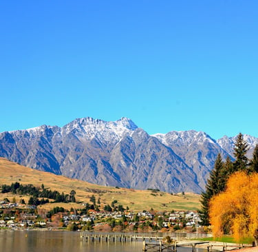 Scenic view of Lake Wakatipu with snow-capped mountains and autumn trees in Queenstown, New Zealand.