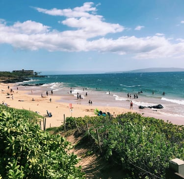 Crowded tropical beach in Maui with people swimming in turquoise ocean water under a blue sky.