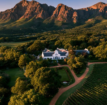 Aerial view of a Cape Dutch manor house and vineyard estate at sunset in Stellenbosch, South Africa.